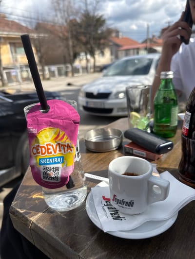 The image presents an outdoor cafe scene in Kumanovo, North Macedonia, under a bright, partially cloudy sky. The foreground shows a dark wooden table with visible grain. On the table, from left to right, there is a clear glass containing water with a pink Cedevita 'Ananas & Mango' powder sachet, marked with a QR code and text in Croatian/Serbian/Macedonian, and a black straw inserted. Adjacent to this is a small white espresso cup, branded "Segafredo Zanetti," holding dark liquid, positioned on a matching white saucer and a folded Segafredo Zanetti napkin. Further right, a silver metallic ashtray is present. An orange-red cigarette pack with a dark grey IQOS device resting on top is also visible. An empty clear glass with a yellowish residue is positioned behind these items, next to a green glass bottle with a silver cap. A dark brown bottle, partially visible on the far right, appears to be a soda. A person, visible from the mid-torso to the head on the right, wears a white short-sleeved shirt and holds a dark smartphone to their right ear, engaged in a phone call. The background reveals a street with a white Skoda sedan parked in the mid-ground. Beyond the street, buildings with light-colored facades and reddish-brown tiled roofs are visible, accompanied by leafless deciduous trees and overhead utility wires.