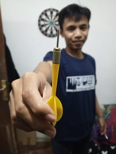 The scene is an indoor setting in a room in Al Haouafate, Morocco. A human hand, positioned in the foreground and in sharp focus, extends a single dart directly towards the viewer. The dart features a silver metal point, a knurled brass-toned barrel, and a bright yellow plastic shaft terminating in three yellow flights. In the blurred background, a male individual with short dark hair and wearing a dark blue t-shirt with a white graphic is partially visible, looking towards the camera with a faint smile. Behind him on a light-colored wall, a traditional dartboard with red, green, black, and white segments is mounted. To the far left, a dark vertical element, possibly a door jamb, with a metallic handle is partially visible.