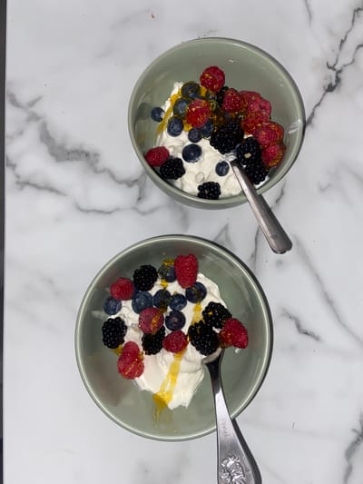 Two light green bowls are set on a white marble surface, each containing a serving of white cream, blueberries, raspberries, and blackberries. A drizzle of honey tops the fruit. A silver spoon rests in each bowl. The location is unknown, but the United Kingdom is requested.