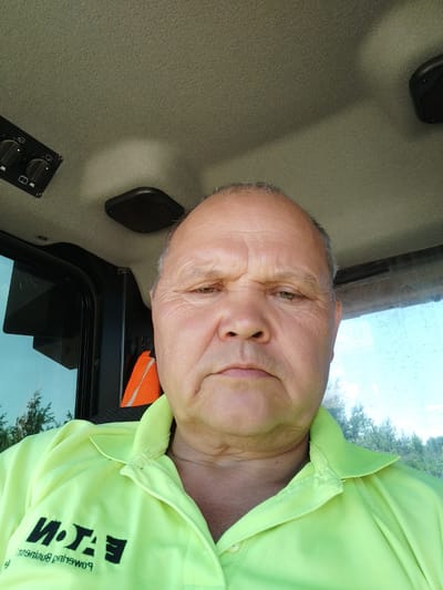 The image shows a middle-aged Caucasian man with fair skin and graying hair, wearing a bright green collared shirt with a black "EATON" logo on the left chest. He appears to be inside a vehicle, possibly a tractor or construction equipment, given the view of the interior ceiling and controls. An orange safety vest is visible behind him. The background through the window shows trees and a bright sky, suggesting an outdoor setting. The man's expression is neutral, and he is looking directly at the camera. The image was taken from a selfie perspective. There is no information in the image that allows for identification of the location as Rozdil, Ukraine.
