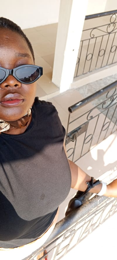 A dark-skinned individual, appearing to be an adult female, is depicted in a high-angle self-portrait photograph. She wears black cat-eye sunglasses that obscure her eyes, a plain black short-sleeved top, and a brown and white patterned scarf around her neck. Her lips exhibit a glossy finish. A small, dark circular mark is visible on the fabric of her top, central to her chest area. On her right wrist, a white band and a black strap or tie are present. The subject is positioned outdoors, facing the camera, with a dark-colored metal railing featuring decorative scrollwork and vertical bars in the midground and foreground to her right. Beyond the railing, a light-colored tiled floor and a light-colored vertical pillar are visible. The scene is illuminated by strong daylight, casting a distinct shadow on the subject's left side. The environment suggests an upper-level outdoor space, such as a balcony or walkway. No specific architectural or geographical markers in the image definitively confirm the location as Ijotan, Nigeria; however, the scene is consistent with a residential or commercial building within an urban setting as specified.