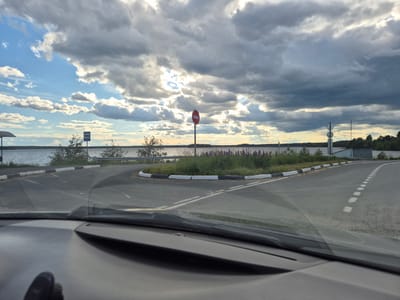 The media file captures a view from inside a vehicle, looking out through the windshield at a road leading towards a body of water. The scene appears to be a rural or semi-rural location in Mashezero, Russia. The road curves to the left, with a distinct black and white curb. A red circular sign with a white horizontal bar, indicating "no entry" or "do not enter," is prominently visible on a pole in the middle distance. To the left of the road, a blue sign with a white bus icon signifies a bus stop.
The setting is a lakeside environment with calm water stretching to the horizon. Lush green grass and bushes, dotted with purple lupine flowers, line the roadside. In the background, a dense line of trees forms the far shore. The sky is dramatic, filled with a mix of dark, heavy clouds and patches of bright blue sky where sunlight is breaking through. This suggests a dynamic weather pattern, possibly transitioning from or towards rain, with a late afternoon or early evening feel due to the lighting. The overall mood is serene, with the natural landscape taking center stage, punctuated by the man-made elements of the road and signs.