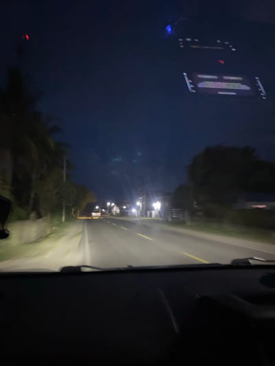 This image depicts a nighttime scene, likely taken from inside a vehicle looking out at a road in Meunasahlhok, Indonesia. The perspective is slightly angled, with the road and its surroundings filling the right side of the frame and the dark sky and reflections dominating the left.
The road is visible with a faint yellow line marking its center, illuminated by artificial lights. Along the side of the road, there are streetlights and the shapes of buildings or houses. The overall atmosphere is dark and somewhat blurry, suggesting movement or low light conditions. Trees and vegetation are visible on the left side of the road, silhouetted against the night sky.
On the left side of the image, there are reflections on a surface, possibly a window. One reflection shows what appears to be a digital display or sign with vertical lines of light in various colors, including purple and white. There is also a small blue light visible in this reflection, and further to the left, a blurred red light can be seen. The dark sky suggests it is nighttime, and no specific weather conditions are apparent beyond the darkness. The scene conveys a sense of travel or passing through an area at night.