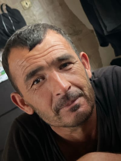 This is a close-up, vertically oriented photograph of a man's face, viewed from a slightly upward angle. The man has dark hair, a beard, and appears to be in his late 30s or early 40s. His forehead is furrowed, creating prominent wrinkles. His eyes are open, with the right eye more visible than the left. He has a mustache and stubble on his chin and jawline. The man is wearing a dark t-shirt, the fabric of which is visible in the upper and lower parts of the image. The background is dimly lit and appears to be an indoor setting, possibly a room with furniture. There are no specific cues about the time of day or weather. The man's expression is difficult to discern definitively, but the furrowed brow might suggest concern or deep thought. No text is visible in the image. Given the location context of Adzhiryuara, Abkhazia, the scene is likely within this region.