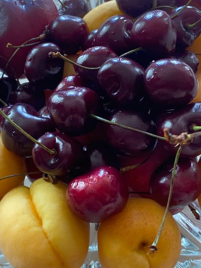 A close-up, overhead shot shows a pile of ripe cherries and apricots in a clear glass bowl. The cherries are a deep red, with some still attached to their stems. The apricots are a vibrant orange-yellow, with a soft blush on one side. The fruits are wet, suggesting they have been recently washed. The background is blurred, but appears to be a light-colored surface. The image does not contain any information about the location in the city of Askarovo, Russia.