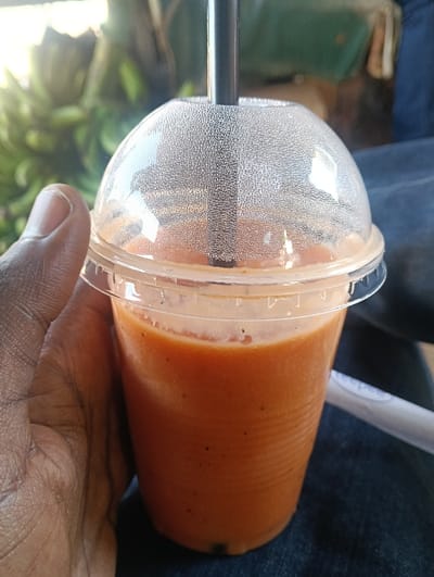 A dark-skinned left hand holds a clear plastic cup filled with an orange-brown liquid, consistent with a blended beverage or smoothie, containing small dark particles. The cup is topped with a clear, textured plastic dome lid, through which a black drinking straw is inserted. The cup is positioned above dark blue fabric, likely denim clothing. In the out-of-focus background, green foliage, including visible bunches of unripe bananas, is illuminated by sunlight. The scene is located in Ndenderu ward, Kenya.