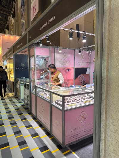 This image depicts an indoor scene at a jewelry exhibition, likely within the Jaipur Exhibition & Convention Centre (JECC) in Jaipur Municipal Corporation, India.
The primary subject is a brightly illuminated "VIBRAANT JEWELS" exhibition booth, constructed with an aluminum frame and pink and white panels. The booth features multiple glass display cases filled with various jewelry pieces, including rings, necklaces, and bracelets, meticulously arranged on stands and trays. These cases are illuminated by internal lights and overhead black track spotlights. The booth's interior walls display large graphics, including a close-up of an eye and a hand wearing a multi-colored gemstone bracelet. Contact information for "SANJAY JAIN (SANSARI)," "HARSHIT JAIN (SANSARI)," and the website "WWW.VIBRAANTJEWELS.COM" is visible on the lower panels.
Inside the "VIBRAANT JEWELS" booth, a male individual with dark hair and a beard, wearing a white shirt, a tan waistcoat, and a lanyard, is leaning forward, actively engaging with the contents of a glass display case. To the left, an adjacent booth, "PM JEWELLER," is partially visible, featuring similar glass display setups in darker tones, with a sign reading "WORLDWIDE MANUFACTURER OF JEWELLERY." A second male individual, dressed in dark clothing, is walking from left to right past the "PM JEWELLER" booth, looking down at a mobile phone held in his left hand.
The floor of the exhibition hall is covered with a patterned carpet featuring alternating rectangular sections of grey, dark grey, and yellow, arranged in a striped configuration. The background reveals the high ceiling and structural elements of the exhibition venue.