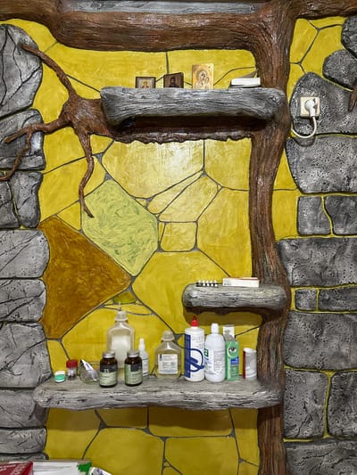 The image shows a shelf structure built into a wall, designed to resemble a tree trunk with branches. The wall behind the shelves is painted with a mosaic-like pattern of yellow and light green geometric shapes. The shelves themselves are made of a textured material that mimics stone or wood.
On the top shelf, there are two small religious icons and a thin, white object, possibly a book or folded paper.
The middle shelf holds a small box and a row of white pills or capsules. A power outlet with a plug and cord is visible on the wall to the right of this shelf.
The bottom shelf is cluttered with various bottles and containers, appearing to be medicinal or personal care products. These include several bottles of liquid, some with labels, and smaller jars or pill bottles. There is also a green box with an eye symbol on it.
The overall impression is of a decorative, custom-built shelving unit in a room with a distinctly artistic or themed wall design. The items on the shelves suggest a personal space, potentially a home or a small clinic, where various personal and medical supplies are stored. The presence of religious icons might indicate a cultural or personal significance to the occupant. There are no people visible in the image. The scene appears to be indoors, given the presence of an electrical outlet. The exact location within Kalininskaya, Russia cannot be determined from the visual information alone.