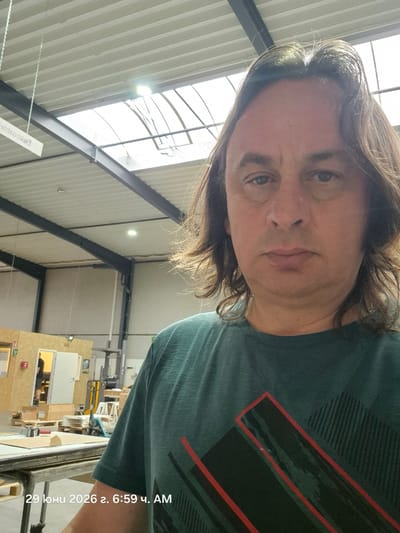 A selfie is taken by a man with long brown hair in what appears to be a warehouse or workshop. He is wearing a dark teal t-shirt with a graphic design featuring red and dark green lines. The background shows industrial shelving, fluorescent lighting, and a skylight. There are some wooden structures and materials visible in the background, suggesting an active workspace. A date and time stamp at the bottom reads "29 Iюни 2026 г. 6:59 ч. AM". The man is looking directly at the camera with a neutral expression.