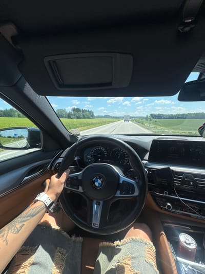 The image shows a first-person perspective from the driver's seat of a BMW car on a sunny day. The driver's hands are on the steering wheel, and they are wearing a white watch and ripped denim shorts. The dashboard displays include a navigation system showing "Ždírec nad Doubravou" and a "Bluetooth: Music Player" screen. Outside the car, a highway stretches into the distance, flanked by green fields and trees under a blue sky with scattered clouds. A white truck is visible on the highway ahead. The car is moving.