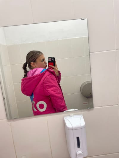 A young female, with her hair in a braid, is taking a selfie in a bathroom mirror. She is wearing a pink jacket over a pink hooded shirt and blue lining. The mirror reflects the white tiled walls and a white soap dispenser mounted on the wall. The overall scene suggests a public restroom.