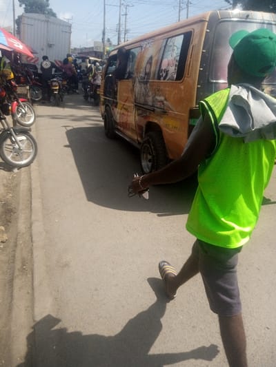 A man in a green vest, cap, shorts, and sandals walks on a sunny street. A yellow van with artwork on its side is parked nearby. Motorcycles line the left side of the road under a market canopy. More motorcycles and people are visible further down the road. Power lines stretch overhead. The scene is shot from a ground-level perspective.