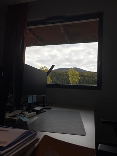 The image depicts an indoor scene looking out through a window to an exterior landscape on an overcast day. The location is Lièpvre, France.

Inside, a white desk occupies the lower half of the frame. On the left side of the desk, a dark computer monitor is visible, facing away from the viewer. A black desk lamp with a curved arm is positioned left of the monitor. Cables and light blue sticky notes are discernible behind the monitor stand. A rectangular grey mousepad is centered on the desk. In the immediate foreground on the right, a stack of documents with blue-bound spines and an orange-brown folder are partially visible. A dark red curtain is partially visible on the far left. The interior is dimly lit.

The exterior view through the dark-framed window reveals an expansive natural landscape under a cloudy, grey sky. A large, forested hill dominates the background. On the crest of this hill, a tall, slender, dark structure, identified as the ruins of Château du Frankenbourg, is clearly visible. In the mid-ground, within the forested area, several trees display prominent bright yellow foliage, indicative of autumn. A dark horizontal band, possibly a road or waterway, lies at the base of the main forested hill. Above the window frame, a section of a wooden slatted ceiling or overhang is visible. No individuals are present in the scene.