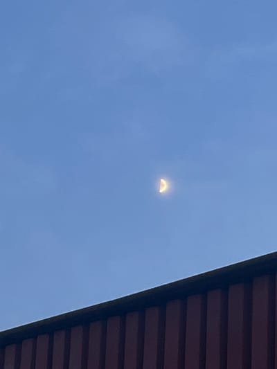The image shows a clear blue sky with a crescent moon visible in the upper right quadrant. In the foreground, the top edge of a dark-colored structure with vertical, reddish-brown elements is visible, extending across the bottom of the frame. The overall scene suggests an outdoor environment, likely at dusk or dawn, given the presence of the moon and the lighting conditions. The location is identified as Ożarów Mazowiecki, Poland.