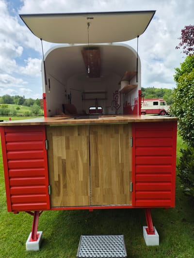 The image shows a red and white mobile bar, likely a converted shipping container, parked on a grassy area. The bar has a wooden countertop, open on one side to reveal the interior. The interior appears to be a food or beverage preparation area with shelves and equipment. A vintage red and white van is partially visible in the background, suggesting a mobile catering or beverage service. The location is on grass in Lenningen, Luxembourg.