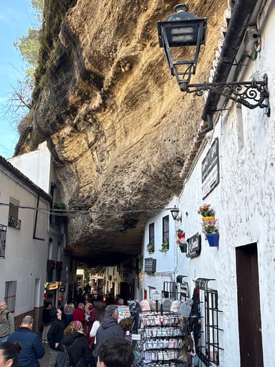 An overhead view captures a busy street in Setenil De Las Bodegas, Spain, characterized by whitewashed buildings constructed directly under and into a massive natural rock overhang. The street runs from the foreground into the mid-distance, where it narrows further and curves slightly.

On the left, multiple multi-story whitewashed buildings with visible red-tiled roofs and small balconies recede into the distance. Power lines crisscross above the street between these structures.

The right side features a prominent whitewashed building whose upper section is directly beneath the large, textured rock overhang. An ornate black streetlamp, featuring a modern LED light panel, is affixed to this building with a scrolled bracket. Below the streetlamp, a section of red-tiled roof gutter is visible, along with a white downspout. Several rectangular signs with dark backgrounds and white text, such as "CARNE Y U..." and "EL CORNETI," are mounted on the facade. Colorful potted flowers in blue, red, and yellow containers are suspended from the wall. The lower portion of this building displays two merchandise racks; one large rack holds numerous hats (beanies and caps) and other small items, while a smaller rack nearby displays additional merchandise. String lights are visible across the building's lower facade.

The dominant geological feature is a massive, sun-drenched rock formation that forms a natural ceiling over the street. The rock exhibits various shades of tan and brown, with visible layers, crevices, and darker streaks of moisture or mineral deposits. Sparse vegetation, including leafless branches and some green foliage, grows from the top edge of this rock face, silhouetted against a clear blue sky in the upper left.

The street is populated by approximately 30-40 individuals, primarily adults, engaged in walking, browsing, and observing the unique environment. They are dressed in casual clothing. Several people are gathered near the merchandise racks on the right, while others move further down the street, which appears to be a pedestrian zone. A vehicle, possibly a white car or van, is partially visible in the distant background, indicating vehicular access or parking deeper within the street.