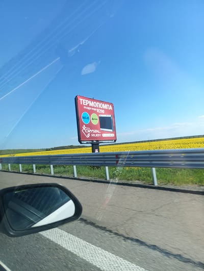 The image, apparently taken from a moving vehicle, shows a roadside scene. The sky is clear and blue, and the road appears to be asphalt. A guardrail runs alongside the road. Beyond the guardrail, a field of yellow flowers is visible.

A large advertising billboard dominates the view. It is predominantly red and features text in Cyrillic, including the words "ТЕРМОПОМПА" and "въздух-вода R290". It also displays "Wi-Fi" and "A+++" inside blue and green circles respectively. Below these are the words "CRYSTAL RUBY". The billboard seems to be advertising a product.

In the foreground of the image, the car's side mirror is visible. The location is somewhere in Vratitsa, Bulgaria, based on the Cyrillic text on the billboard.
