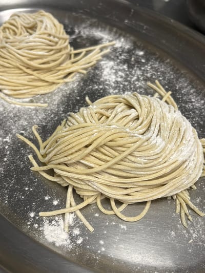 The image displays two bundles of raw, light-beige pasta noodles, each coiled into a nest, resting on a metallic surface in a food preparation area in Pozuelo de Alarcón, Spain. Both noodle bundles are uniformly coated with white flour. The larger bundle occupies the foreground, in sharp focus, while a smaller, similarly prepared bundle is visible in the upper background. The metallic surface itself is dusted with flour, particularly around the pasta, and exhibits a subtle brushed texture. No individuals are present in the scene.