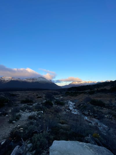 The image depicts a landscape scene in Unknown, Argentina. The sky is clear blue, taking up a significant portion of the frame. Adjacent to the sky is a mountain range. On the right side of the image is a steep, rocky terrain, with sparse vegetation. The foreground consists of a rocky cliff. The overall tone of the image suggests a natural, rugged environment.