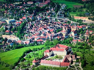This is an aerial photograph of the city of Ellwangen, Germany. The scene captures a dense urban landscape interspersed with lush green fields and forests. The architecture is characteristic of European towns, with many buildings featuring red-tiled roofs. A prominent feature in the foreground is a large, sprawling complex of light-colored buildings with red roofs, likely a historical site or institution. To the left of this complex, a circular structure with battlements suggests a fortification or old defensive structure. Further into the background, the town extends uphill, showcasing a dense collection of houses and buildings, some with spires, hinting at churches or prominent landmarks. Roads and pathways weave through the town and surrounding landscape. The time of day appears to be daytime, with bright sunlight casting shadows, indicating fair weather. The overall impression is one of a well-established, historic European town nestled within a verdant, natural environment. There is no visible text in the image.