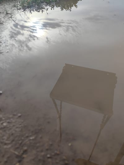 The image shows a reflection in a puddle, depicting the sky, some vegetation, and a partially submerged object that appears to be a small, square table with decorative beaded edges. The reflection of the sky is bright, suggesting it is daytime, with clouds and the sun visible. The vegetation at the top of the image consists of green leaves and stems, hinting at an outdoor setting, possibly near a body of water or in a damp area. The table's reflection is dark and indistinct, suggesting it is not fully illuminated or is made of a dark material. The water is murky, with a sandy or muddy bottom visible in some parts of the reflection, suggesting a natural outdoor environment. The overall mood is somewhat abstract and contemplative, with the reflection creating a distorted or layered reality. The location is identified as Dolan, Kazakhstan.