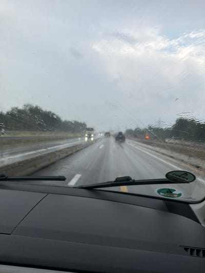 The image shows a view from inside a vehicle looking out through a rain-streaked windshield onto a highway in Weinheim, Germany. It is daytime, and the weather is overcast with rain. Several cars are visible on the highway, some with their headlights on. The road is wet, and trees line the sides of the highway. There are two circular stickers on the windshield, one of which appears to be from DEKRA. The dashboard of the vehicle is visible in the foreground.