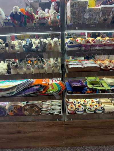 This image shows a display of traditional Kyrgyz handicrafts in a market stall located in Bishkek, Kyrgyzstan. The stall features several shelves filled with various handmade items. On the top shelf, there are several plush dolls dressed in traditional clothing, some with animal motifs. Below these, another shelf holds a collection of small, decorative yurt-shaped items, each adorned with colorful patterns and traditional designs. Further down, a shelf displays intricately embroidered fabric items, possibly placemats or coasters, in vibrant colors and patterns. Another shelf showcases an assortment of felt hats with decorative embroidery. The overall scene is one of a vibrant and well-stocked market stall offering a variety of cultural artifacts for sale.