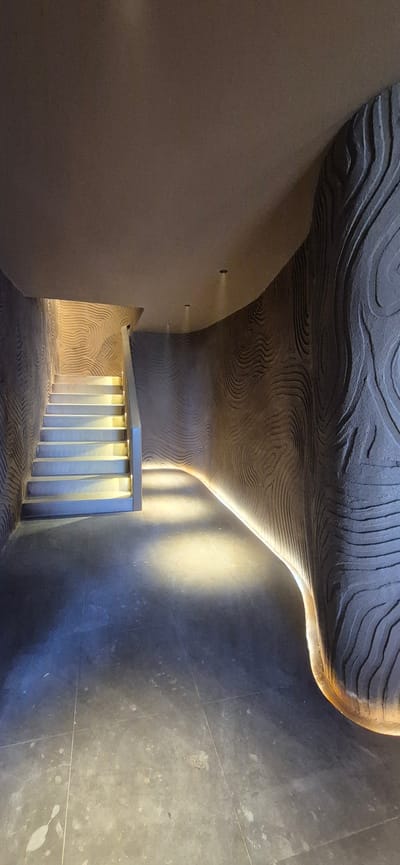 The image shows a concrete staircase with illuminated risers and a railing, leading upwards into a dimly lit area. The walls on either side of the staircase and extending into the background are textured with organic, wave-like patterns and are illuminated by a warm-toned LED strip light running along the base of the curved wall. The floor is made of dark, tiled material. The scene appears to be an indoor architectural space, possibly a modern building or a tunnel. The style suggests it could be located in Ashgabat, Turkmenistan, given the country's penchant for futuristic and unique architectural designs, particularly those incorporating curved forms and extensive use of concrete and lighting. There are no people visible in the image.