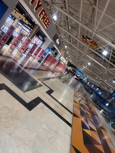 The image shows the interior of what appears to be an airport terminal in Addis Ababa, Ethiopia. The perspective is skewed, taken from a low angle looking down a long corridor. The ceiling is high with an exposed framework of white metal beams and visible light fixtures. A large, brightly colored abstract art piece hangs from the ceiling.

On the left side of the corridor are duty-free shops with prominent signs such as "DUTY FREE", "GUERLAIN", and "LANCÔME". There are displays of products within the shops, and signs in red and white are visible along the storefronts.

People can be seen in the distance, moving through the terminal. There are also signs indicating gate information, specifically "A8-A19" and "B, C & D."

The floor has a patterned design, with large sections of marble-like tiles accented by black zigzagging inlays, leading to a geometric, triangular design closer to where the terminal gates are. There's a yellow triangle design next to the terminal seating area.