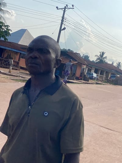 This image captures an outdoor scene in Igbara, Nigeria, under a bright, partly cloudy sky.

In the foreground, slightly to the left, a man of dark complexion is visible from the chest up, facing right with a neutral expression. He has a bald or closely shaved head and wears a dark olive-green polo shirt with a black collar and visible buttons. A small, circular, glowing white emblem is affixed to the left side of his chest.

The midground features a reddish-brown unpaved road or path. Further back on the right, another individual, dressed in a patterned purple and white garment, walks away from the viewer along a dirt path adjacent to several single-story buildings. These buildings have various colored walls, often with rusted corrugated metal roofs, and some feature verandas. Two cars, one light blue, are parked near these structures.

In the background, to the left, a distinctive white, pyramid-shaped building with a small cross-like structure at its apex is prominent against the sky. To its right, a wooden utility pole with multiple cross-arms and numerous overhead power lines diagonally spans the upper right quadrant of the image. A street light is attached to the pole. Sparse green foliage, including palm trees on the right, is scattered throughout the scene. The sky is a light blue with scattered white cumulus clouds.