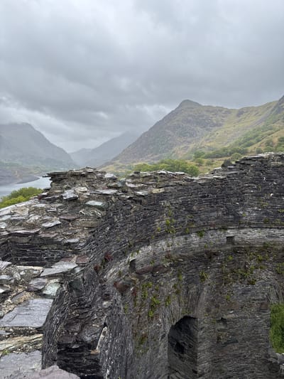 The image depicts a stone structure, possibly the ruins of a building or fortress, situated in a mountainous landscape on a misty day, most likely located near Llanberis, United Kingdom. The structure appears to be made of dark, layered stone, and there is an arched opening or hole in the wall. The landscape features green, sloping hills or mountains in the background, shrouded in low-hanging clouds or mist. The overall environment appears rugged and natural, with an overcast sky contributing to a subdued lighting.