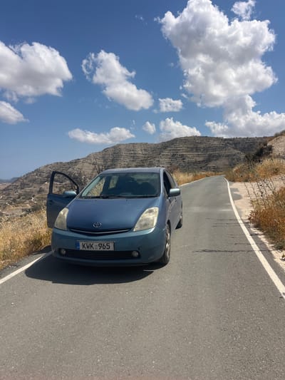 A blue Toyota Prius is parked on the side of a paved road in Agios Georgios Lemesou, Cyprus. The car's driver-side door is open. The road curves to the right and ascends a hill. The landscape is arid with dry grass and sparse vegetation on either side of the road. In the background, a barren hillside is terraced with numerous horizontal lines, suggesting past agricultural use. The sky is blue with scattered white clouds. The license plate of the car reads "KWK 965".