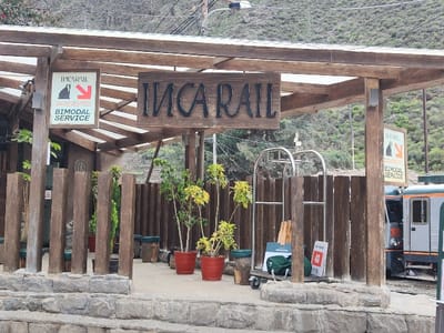 The image shows an Inca Rail train station with its signage. The station is covered by a wooden pergola, with a sign reading "INCA RAIL" in dark lettering. Below, a wooden fence-like barrier surrounds the station. Several potted plants and a luggage cart are visible. To the right, a silver and orange train is partially seen. The station appears to be situated in a mountainous area, with trees and hills visible in the background.