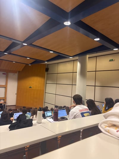 The image depicts a modern lecture hall or auditorium, likely located in Porto Salvo, Portugal. The perspective is from the mid-rear section of the room, looking towards the front.

The ceiling features a grid design composed of dark blue beams intersecting light-brown, wood-paneled squares. Numerous recessed circular lights illuminate the space; several bright lights in the foreground ceiling create glare.

The left wall in the background is clad with tall, light-brown wood panels, featuring a green emergency exit sign and a red "STOP" sign. A screen or whiteboard is partially visible on the far left. The right wall in the background consists of light-colored, segmented panels arranged in a grid pattern, also displaying a green emergency exit sign. A large, cylindrical white column stands centrally on the right side of the room, extending from floor to ceiling.

A large group of individuals, primarily appearing to be students, are seated in tiered rows of light-colored desks, facing forward. Most individuals are seen from behind or the side. Many have open laptop computers on their desks, displaying various screens.

In the immediate midground, a male subject wearing a white t-shirt with a colorful graphic on the back is seated, facing away from the camera. His right arm is bent, with his hand near his head. To his left, a female subject with long dark hair is also focused on a laptop. To his right, another female with long dark hair wears a yellow garment.

Objects on the desks in the midground include a white insulated water bottle and a transparent purple bottle containing a beverage, possibly with berries. Papers and notebooks are visible. A black jacket or fabric item is draped over a seat or desk to the left. In the extreme foreground on the right, a folded white fabric item with a colored pattern is partially visible. The overall scene suggests an academic setting during a lecture or study session.