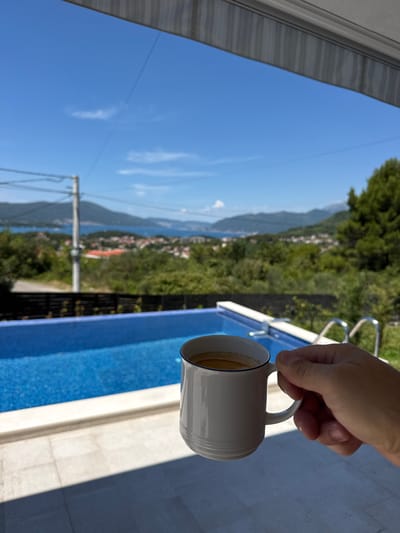 A hand holds a white mug of coffee in the foreground, with a blue swimming pool and a coastal town in the background. The scene appears to be a residential area in Kavač, Montenegro. The mug is white with a thin blue rim, and the coffee is a light brown color. The swimming pool is rectangular with a tiled edge. In the distance, there are buildings with orange roofs, lush green trees, and mountains. The sky is clear blue with a few wispy clouds. The overall impression is one of a peaceful and scenic morning.