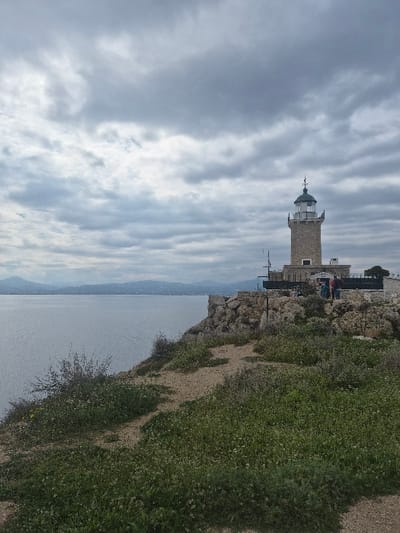 An image depicts a coastal scene at Cape Heraion in the Municipal Unit of Loutraki - Perachora, Greece. A stone lighthouse with a white lantern room and a dark green dome stands prominently on a rocky promontory on the right side of the frame, positioned above a body of water. Approximately four to five individuals are observed standing near the base of the lighthouse, partially obscured by the structure and rocks. The immediate foreground features uneven, sandy-brown terrain interspersed with patches of green vegetation, including small shrubs and what appear to be wildflowers. A calm, gray-blue body of water occupies the left and central midground, extending to a distant, hazy landmass of hills or mountains under an overcast sky filled with numerous gray and white clouds. The overall lighting suggests a muted daylight condition.