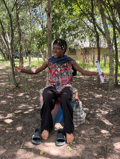 An individual is seated outdoors in Chitungwiza, Zimbabwe, within a partially shaded, natural environment. The person, with dark skin and dreadlocked hair adorned with colorful beads, is wearing a sleeveless, multi-patterned top with geometric and abstract designs in red, blue, yellow, and black, a dark blue scarf around the neck, and black trousers. A silver-colored pendant is visible on the shirt. The individual is positioned on a painted drum or barrel, exhibiting red, blue, and white sections. Both arms are outstretched; the left hand is open, and the right hand holds a white rolled document. The facial expression conveys animation, with an open mouth suggesting speaking, laughing, or singing. Two black slide sandals are placed on the dirt ground directly in front of the individual's feet, which wear orange-colored socks. A plaid fabric bag is situated on the ground to the individual's left. The ground is dirt, covered with scattered dry leaves, small branches, and sparse green vegetation. The scene is illuminated by bright daylight, casting dappled shadows. In the background, numerous trees with green foliage are visible. To the right, a light-colored brick building with a thatched roof and dark-framed windows is partially obscured by trees. To the far left, a teal-colored pickup truck is partially visible through the trees.