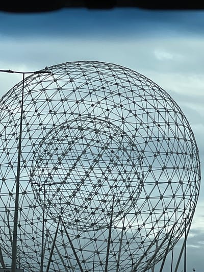 An outdoor, medium shot captures a large, geodesic sphere sculpture against a cloudy sky. The sculpture is constructed from a complex network of intersecting metal rods forming triangular patterns, with a smaller, concentric sphere visible within the larger one. A street light pole stands to the left of the sculpture, and a dark, horizontal band at the top of the image suggests a vehicle's windshield. The sculpture is likely located in Belfast, Northern Ireland, United Kingdom, though no specific landmarks are identifiable in the frame.