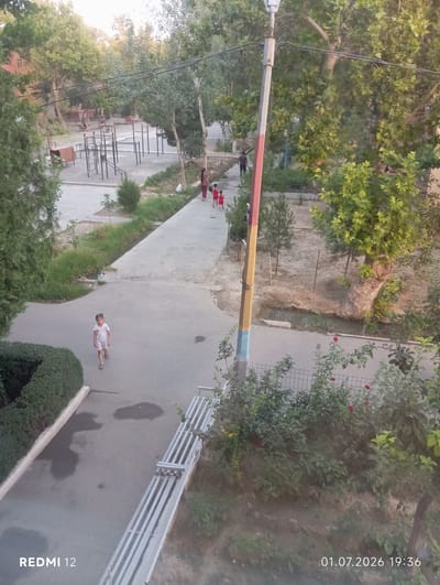 This is an outdoor, eye-level shot of a scene in Bekabad, Uzbekistan. The photograph is oriented vertically. The subject of the image is a paved pathway winding through a park-like setting. A young child, dressed in a white outfit, is walking along the pathway. In the background, there is a playground with various metal exercise equipment. Mature trees with lush green foliage surround the area, casting dappled shadows. The lighting suggests it is late afternoon or early evening, with a warm, soft glow. The weather appears to be pleasant. The pathway is bordered by grass and bushes, with some areas of bare earth. In the upper right corner of the image, text reads "01.07.2026 19:36". Along the bottom right edge of the image, text reads "REDMI 12".