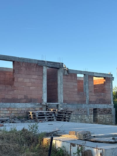 This is a ground-level, medium-shot, vertical, outdoor, daytime photograph taken in Enevo, Bulgaria. The scene depicts a partially constructed building made of red brick and concrete. The structure has large, rectangular openings where windows or doorways will eventually be. Exposed rebar extends from the top of the concrete pillars. Piles of wooden pallets are stacked in front of the brick wall, and various construction debris, including wood planks and broken bricks, are scattered on the ground. The foreground shows a concrete slab with some overgrowth of dry grass and small plants. In the upper portion of the image, a clear blue sky is visible with a thin white contrail from an aircraft. The lighting suggests it is early morning or late afternoon, casting shadows on the building and ground. There are no people visible in the frame.