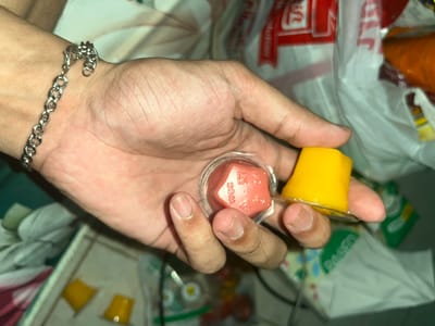A hand holds two small, round, clear plastic containers, each with a foil lid. The container on the left holds a bright yellow, gelatinous substance, possibly a dessert or drink. The container on the right holds a reddish-pink, gelatinous substance with a distinct geometric shape inside, resembling a dice. The dice appears to have some markings on its faces, including the letters "WCC." The hand is positioned over a blurry background of plastic bags, one of which has red and white branding. The lighting suggests an indoor setting. The overall impression is a casual, everyday scene.