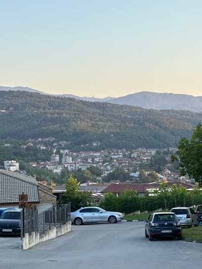 The image depicts a scenic overlook of the city of Kerenite, Bulgaria, characterized by its mountainous backdrop and dense residential areas. In the foreground, a paved road leads downhill, with several parked cars lining its side. Among them, a dark-colored Volkswagen Golf with license plate BT 8232 MA is prominently featured, alongside a silver sedan and a black Porsche with license plate CB 2200 KE. A fence separates the road from a building with a tiled roof and a stone facade. A "P" sign with an arrow indicating parking is affixed to the building. Beyond the immediate foreground, the city unfolds, with buildings clustered on the slopes of a verdant, tree-covered mountain range that dominates the background. The sky above is a clear, pale blue, transitioning to a warmer, yellowish hue near the horizon, suggesting either early morning or late afternoon. The overall impression is one of a tranquil, rural-urban landscape.
