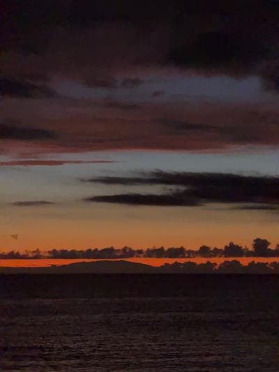 The image displays a close-up view of a reflective surface with a dark, textured background. The reflective surface shows distorted reflections of what appears to be a sunset or sunrise, with colors ranging from deep oranges and reds to lighter yellows and grays. Vertical streaks of darker colors, possibly black or dark brown, are also visible within the reflection, suggesting objects or shadows. On the far right of the image, a dark, wood-like texture is apparent, possibly the surface behind the reflective material. The overall impression is abstract, focusing on the play of light and color on a curved or uneven surface. There is no visible text. The location context of Adeje, Spain, might suggest a coastal sunset, but the image itself does not provide direct visual cues to confirm this. The time of day is implied by the sunset colors, and the weather is likely clear to allow for such vibrant reflections. No people or specific objects are clearly identifiable within the reflection due to the distortion. The emotions evoked are contemplative and perhaps awe-inspiring, due to the dramatic colors and abstract nature of the image.
