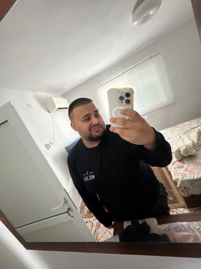 A man is taking a mirror selfie in a bedroom. He is wearing a black long-sleeved shirt and holding a white iPhone in his right hand, with his left hand resting on the phone. The man is looking at his reflection, with his head tilted slightly. Behind him, a bed with a patterned duvet is visible. To the left of the bed, a mirror reflects the room, including an air conditioner mounted on the wall. The room appears to be in the city of Byala Reka, Bulgaria.