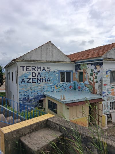 The image shows a building in Soure, Portugal, identified by a vertical mosaic that reads "TERMAS DA AZENHA". The building has a light-colored facade with tiled mosaics depicting different scenes, including a tree and water motif, and colorful fish and dragonflies. The building has windows with light blue trim. A red-tiled roof is visible. Adjacent to the building, there's a concrete wall with grass growing. Blue metal railings and steps are in front of the building. The sky above is overcast with many clouds.