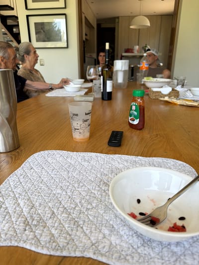 A high-angle shot of a dining table with a white quilted table runner and an empty bowl with a spoon. Beside the bowl are a remote, a bottle of sauce, and a white plastic cup with cartoon drawings. Further down the table are a wine bottle, glass, and multiple white bowls. Two elderly people and a younger person are visible at the end of the table.