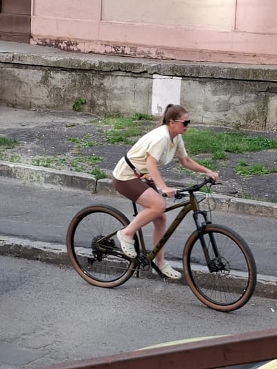 A person is riding a bicycle on a street in Меткомелино, Russia. The person is wearing a light-colored t-shirt, brown shorts, sunglasses, and white clogs. They are in the process of performing a wheelie. The bicycle is dark green with tan tires. The background consists of a textured concrete wall on the left, a grassy area with some weeds, and a concrete sidewalk and street. The lighting suggests it is daytime, likely overcast given the absence of strong shadows. The overall scene appears to be an urban or semi-urban environment. No visible text is present in the image.