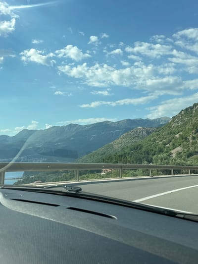 The image captures a scenic view from a vehicle, likely on a highway or coastal road, overlooking a dramatic mountainous landscape. The scene is dominated by towering, verdant mountains that recede into the distance, their slopes covered in lush green vegetation. The sky is a vibrant blue, dotted with fluffy white clouds that are characteristic of a clear, sunny day.
In the foreground, a portion of the car's interior is visible, including the dashboard and a section of the windshield. A metal guardrail runs alongside the road, separating it from the steep drop-off towards the mountains and possibly a body of water below, though it is not clearly visible. The road itself appears to be paved and has a white line marking its lane. A small building with a red roof is nestled on the mountainside in the mid-ground, offering a hint of human presence in the otherwise wild terrain.
The perspective suggests the photograph was taken while the car was in motion, looking out towards the expansive natural beauty. The lighting indicates it is daytime, likely mid-morning or afternoon, with the sun casting natural light on the landscape. The overall mood is one of awe and tranquility, highlighting the grandeur of nature. The location context provided, Blizikuće, Montenegro, aligns with the rugged coastal mountain scenery often found in that region. There is no visible text in the image.