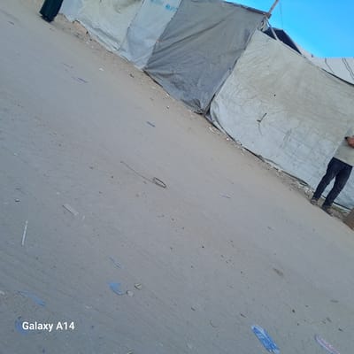 The image depicts a sandy area in Deir el-Balah, Palestinian Territories, characterized by a series of large, tarp-like structures, possibly temporary shelters. The tarps are predominantly white and gray, with some showing signs of wear and tear. One tarp has the UNICEF logo visible. In the foreground, the sandy ground is scattered with debris, including small pieces of plastic and what appears to be a fallen rope or cord with a loop. On the left side of the image, a person in a dark green garment is partially visible, walking away from the camera. On the right side, a person wearing dark jeans and sandals stands with their arms crossed, leaning against one of the tarp structures. The sky above is clear and blue. The overall scene suggests a settlement or temporary living arrangement in a dry, possibly arid environment. The bottom left corner of the image shows a watermark indicating "Galaxy A14".