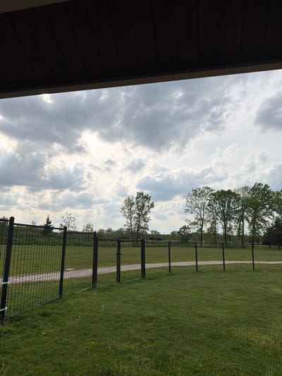The image captures a view of a grassy field bordered by a black metal fence in Glenview, Illinois, USA. A gravel path runs parallel to the fence, visible through the mesh. Trees are interspersed along the fence line, their foliage adding a touch of green against the cloudy sky. The sky is predominantly overcast, with patches of bright sunlight breaking through. In the foreground, the edge of a wooden structure, likely a building or shelter, is visible, providing a sense of context to the scene. The composition suggests a park or recreational area.